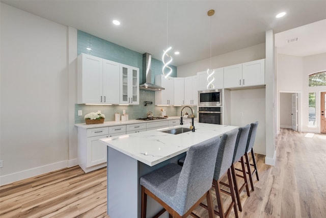 kitchen with white cabinetry, sink, wall chimney range hood, stainless steel appliances, and a center island with sink