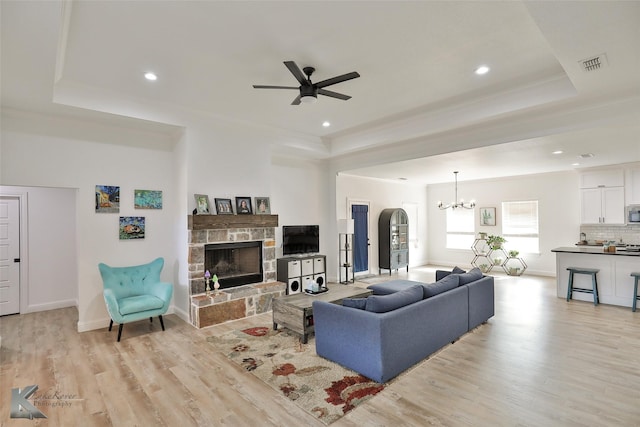 living room with a tray ceiling, a stone fireplace, light hardwood / wood-style floors, and ceiling fan with notable chandelier