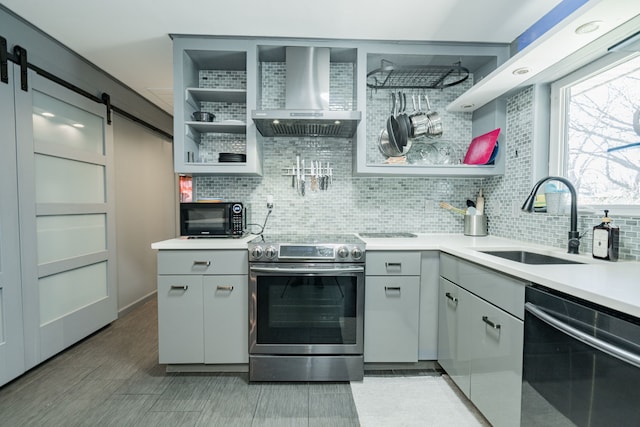 kitchen with gray cabinetry, sink, wall chimney exhaust hood, a barn door, and appliances with stainless steel finishes