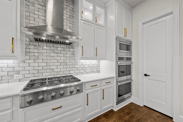 kitchen with appliances with stainless steel finishes, white cabinetry, backsplash, dark wood-type flooring, and wall chimney exhaust hood