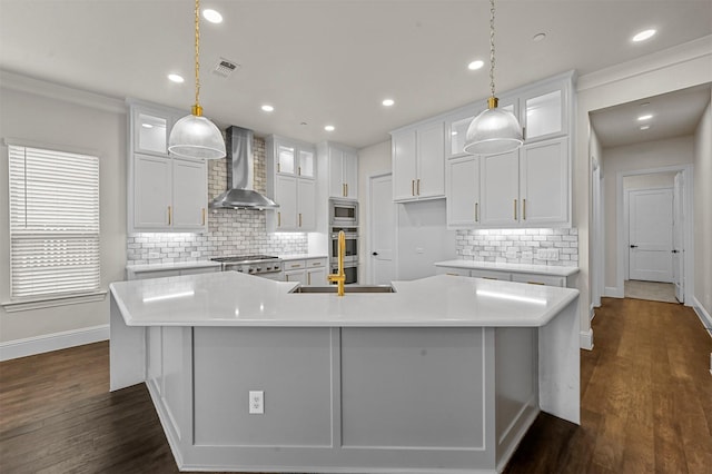 kitchen featuring pendant lighting, appliances with stainless steel finishes, white cabinetry, wall chimney exhaust hood, and a large island with sink