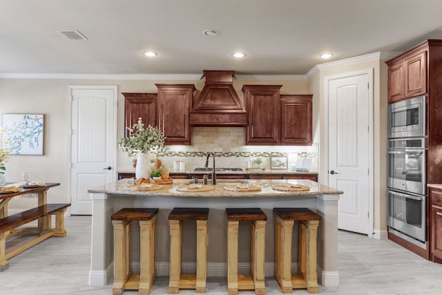 kitchen featuring custom exhaust hood, a kitchen island with sink, light stone counters, a breakfast bar area, and stainless steel appliances