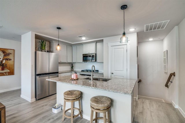 kitchen featuring appliances with stainless steel finishes, gray cabinetry, light stone counters, an island with sink, and decorative light fixtures