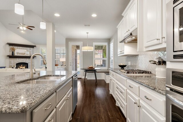 kitchen featuring a stone fireplace, decorative light fixtures, an island with sink, sink, and white cabinets