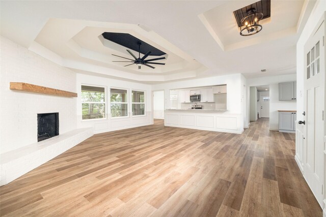 unfurnished living room featuring a raised ceiling, light wood-type flooring, ceiling fan with notable chandelier, and a brick fireplace