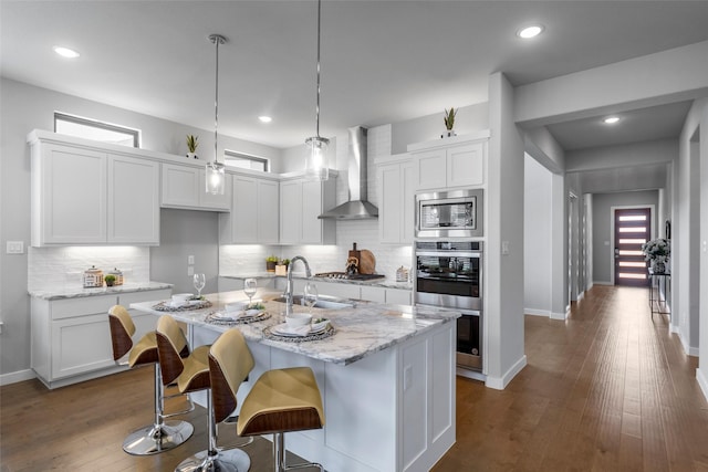 kitchen with white cabinetry, an island with sink, wall chimney range hood, and appliances with stainless steel finishes