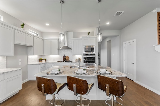 kitchen with sink, wall chimney exhaust hood, hanging light fixtures, an island with sink, and appliances with stainless steel finishes