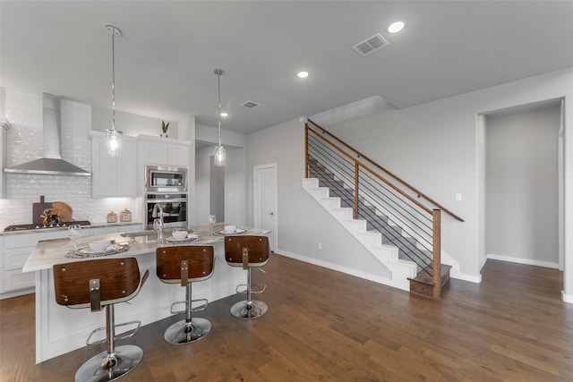 kitchen with appliances with stainless steel finishes, wall chimney range hood, pendant lighting, a center island with sink, and white cabinetry