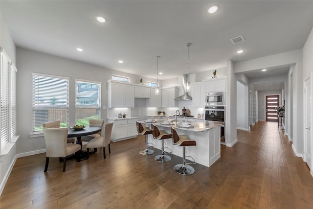 kitchen featuring pendant lighting, wall chimney range hood, an island with sink, white cabinetry, and stainless steel appliances