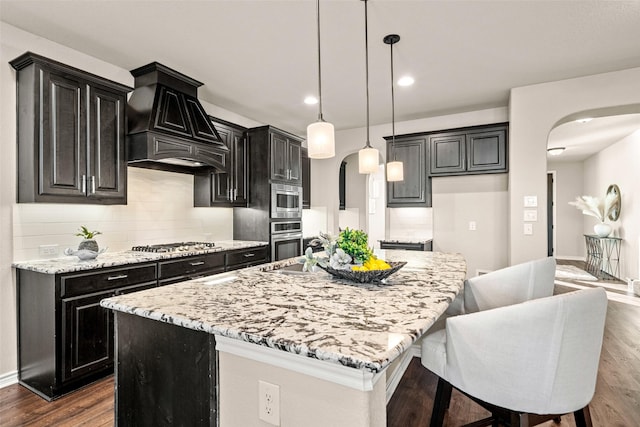 kitchen with stainless steel appliances, hanging light fixtures, light stone counters, a center island with sink, and custom exhaust hood