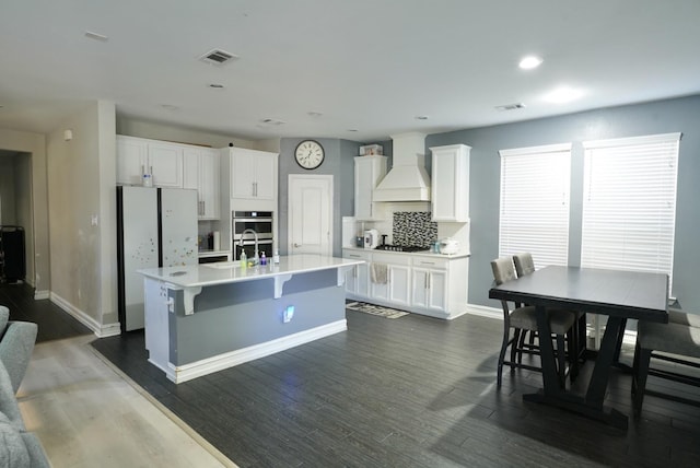 kitchen featuring a breakfast bar area, a kitchen island with sink, stainless steel appliances, custom range hood, and white cabinets