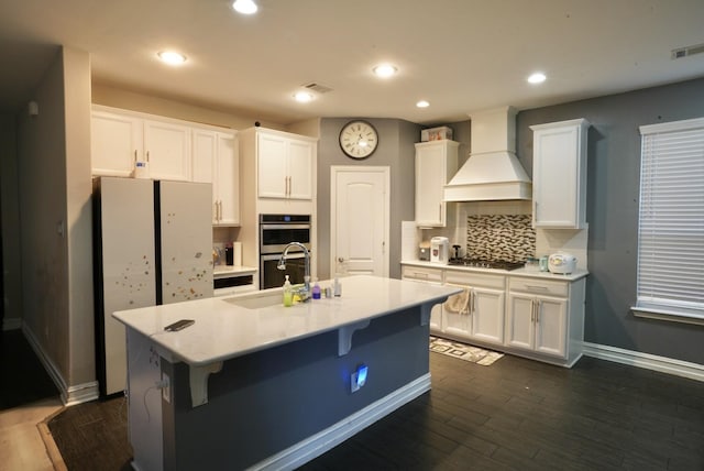 kitchen featuring premium range hood, a kitchen island with sink, sink, and white cabinets