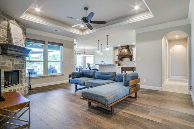 living room featuring a stone fireplace, crown molding, a raised ceiling, hardwood / wood-style flooring, and ceiling fan with notable chandelier