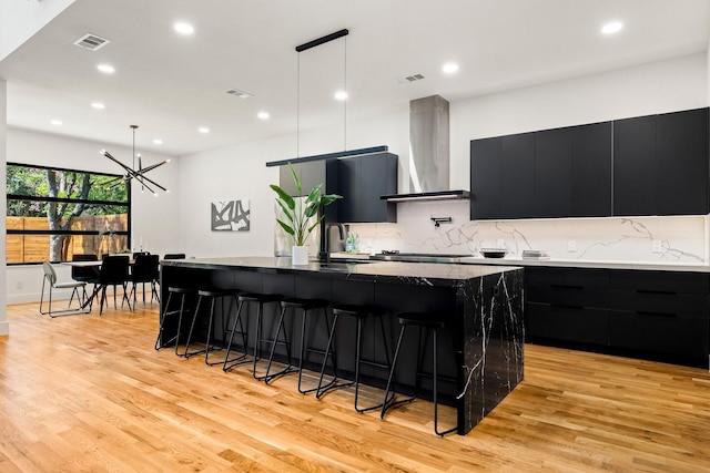 kitchen featuring decorative backsplash, wall chimney range hood, a large island with sink, light hardwood / wood-style floors, and hanging light fixtures