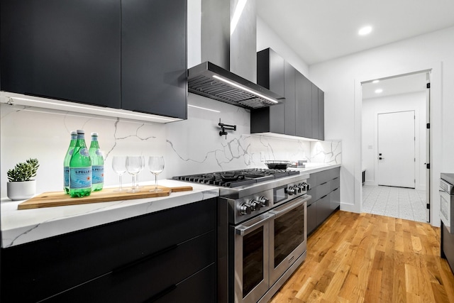 kitchen featuring double oven range, wall chimney range hood, decorative backsplash, light stone countertops, and light wood-type flooring