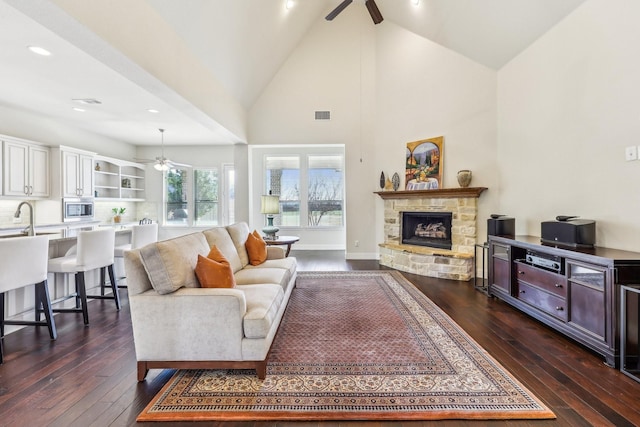 living room with ceiling fan, sink, high vaulted ceiling, dark hardwood / wood-style floors, and a stone fireplace