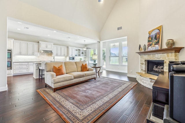 living room with ceiling fan, a stone fireplace, dark wood-type flooring, and high vaulted ceiling