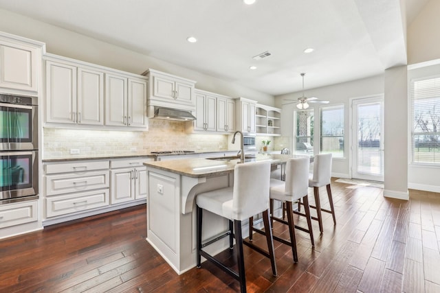 kitchen featuring stainless steel double oven, ventilation hood, a kitchen island with sink, sink, and pendant lighting