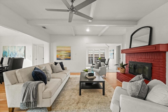 living room featuring beam ceiling, light hardwood / wood-style floors, a brick fireplace, and ceiling fan