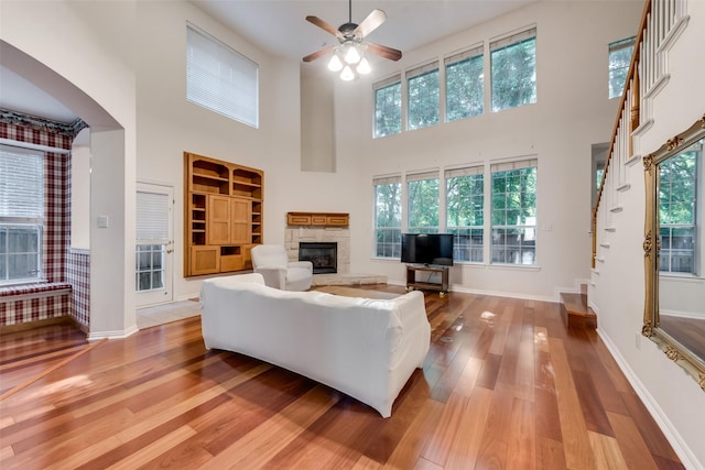 living room featuring wood-type flooring, a towering ceiling, a stone fireplace, and ceiling fan