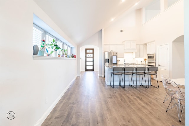 interior space with a breakfast bar, high vaulted ceiling, white cabinets, a center island with sink, and appliances with stainless steel finishes