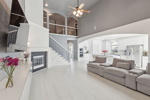 living room with a tiled fireplace, a towering ceiling, ceiling fan, and light wood-type flooring