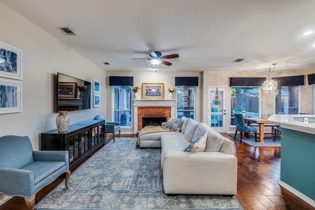 living room with ceiling fan with notable chandelier, dark wood-type flooring, and a brick fireplace