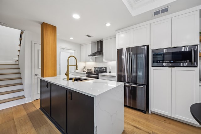 kitchen with a center island with sink, stainless steel appliances, white cabinetry, and wall chimney range hood