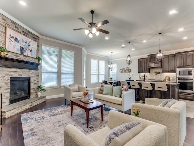 living area with ceiling fan with notable chandelier, a stone fireplace, dark wood-type flooring, and crown molding