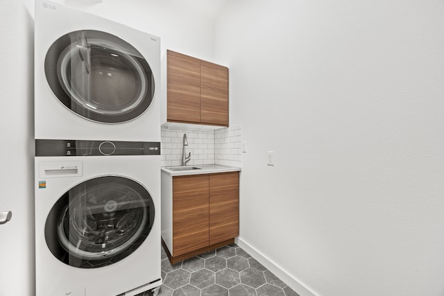 laundry area featuring cabinet space, baseboards, stacked washer and clothes dryer, dark tile patterned floors, and a sink