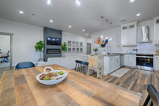 kitchen featuring wall chimney exhaust hood, hanging light fixtures, a fireplace, white cabinets, and appliances with stainless steel finishes