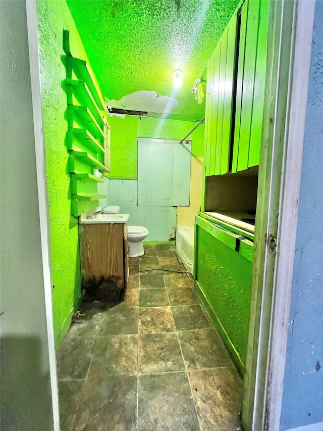 bathroom with shower / bathtub combination, a textured wall, toilet, stone finish floor, and vanity