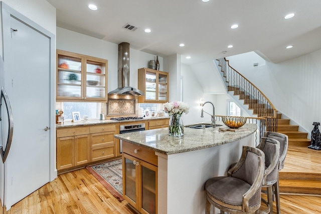 kitchen with light stone countertops, decorative backsplash, wall chimney range hood, light hardwood / wood-style flooring, and an island with sink