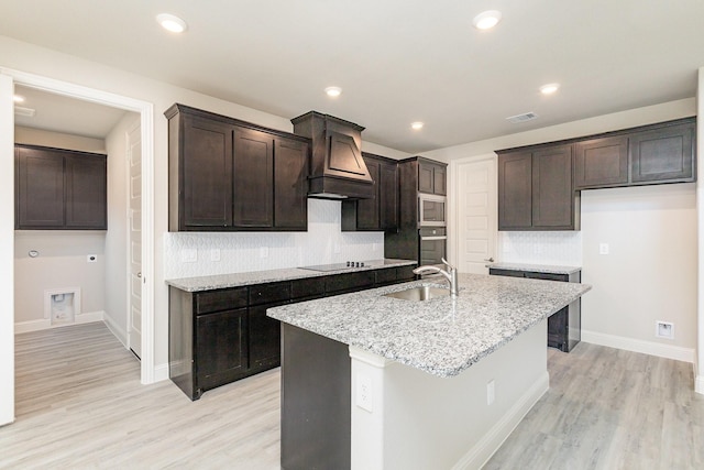 kitchen with light stone countertops, stainless steel microwave, sink, an island with sink, and light wood-type flooring