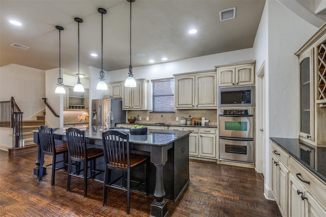 kitchen with decorative backsplash, stainless steel appliances, a kitchen island with sink, cream cabinetry, and hanging light fixtures