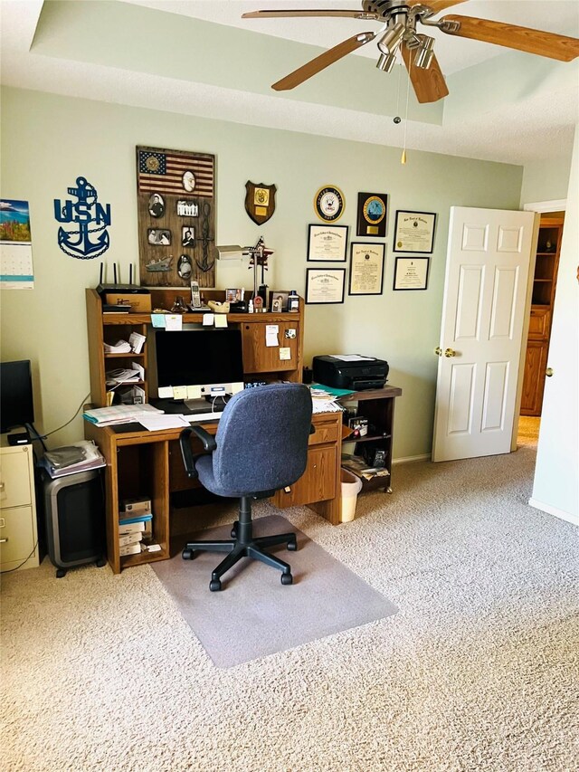 carpeted home office with ceiling fan and a tray ceiling