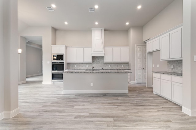 kitchen featuring built in microwave, dark stone countertops, an island with sink, white cabinets, and custom exhaust hood