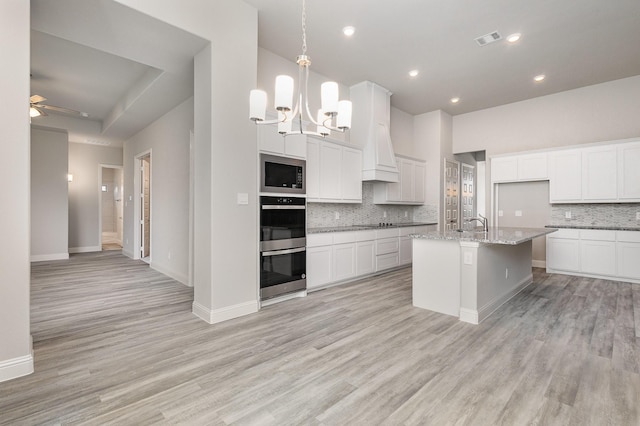 kitchen featuring light stone countertops, ceiling fan with notable chandelier, built in microwave, decorative light fixtures, and an island with sink