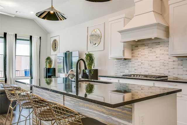 kitchen with dark stone counters, a center island with sink, white cabinets, custom range hood, and appliances with stainless steel finishes