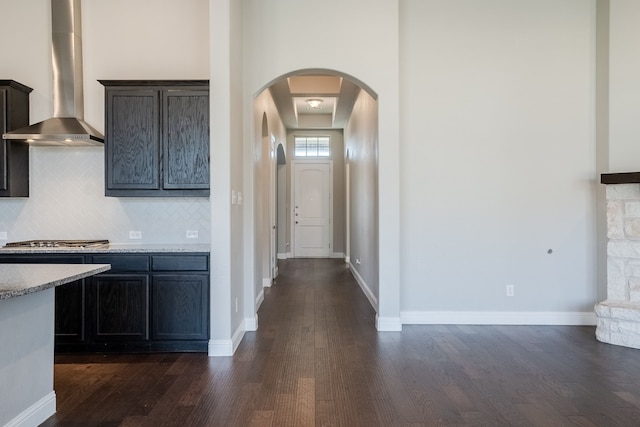 kitchen featuring dark wood-type flooring, wall chimney range hood, a stone fireplace, stainless steel gas stovetop, and decorative backsplash