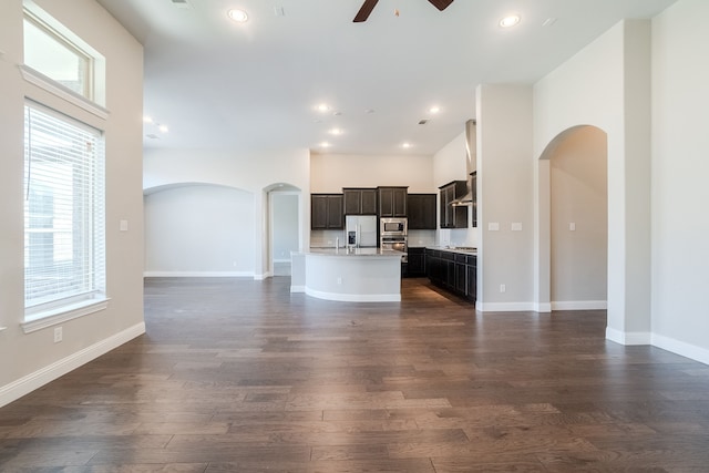 kitchen featuring ceiling fan, wall chimney range hood, dark hardwood / wood-style floors, an island with sink, and appliances with stainless steel finishes