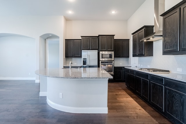 kitchen featuring backsplash, dark wood-type flooring, a center island with sink, wall chimney exhaust hood, and appliances with stainless steel finishes