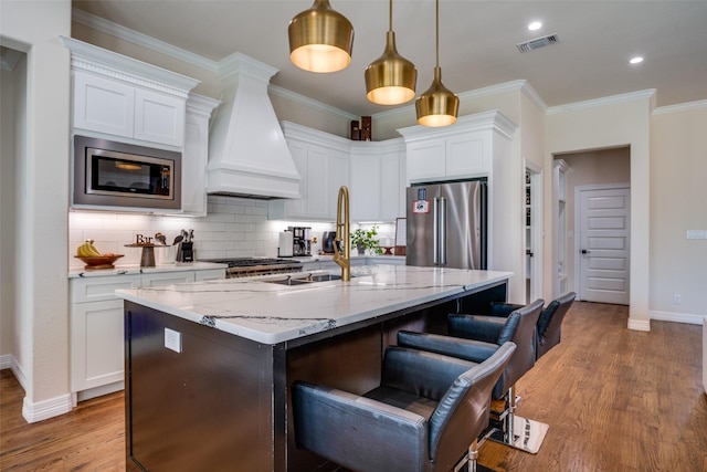 kitchen featuring custom range hood, stainless steel appliances, decorative light fixtures, a center island with sink, and white cabinetry