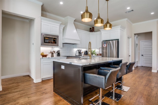 kitchen featuring light stone countertops, custom range hood, stainless steel appliances, white cabinetry, and an island with sink