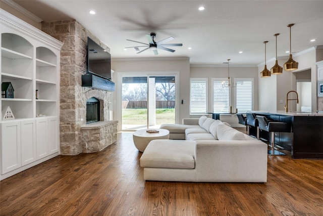 living room with ornamental molding, built in shelves, ceiling fan with notable chandelier, dark hardwood / wood-style floors, and a stone fireplace