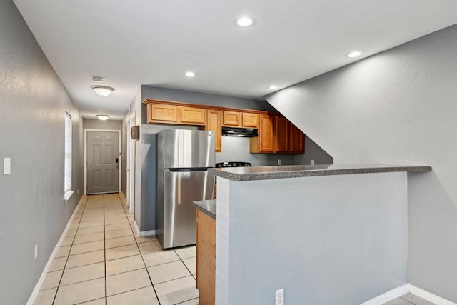 kitchen with brown cabinetry, dark countertops, freestanding refrigerator, a peninsula, and under cabinet range hood