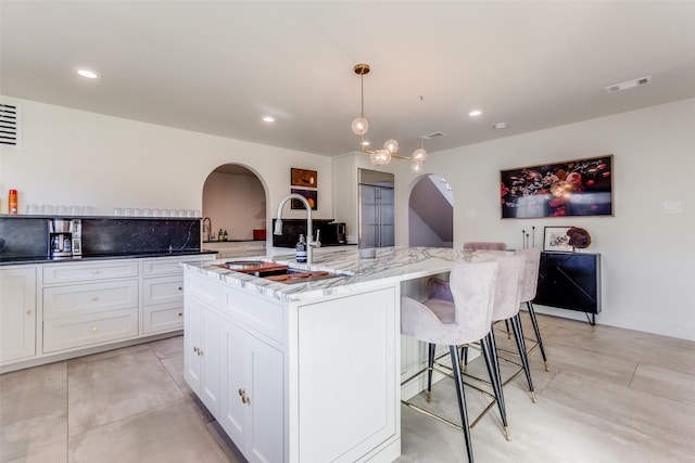 kitchen featuring a kitchen bar, light stone counters, a kitchen island with sink, decorative light fixtures, and white cabinetry