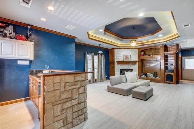 interior space with ornamental molding, a raised ceiling, ceiling fan, light hardwood / wood-style flooring, and a stone fireplace