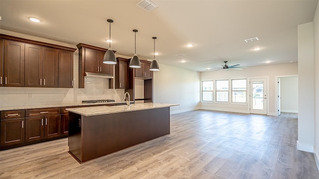 kitchen featuring hanging light fixtures, tasteful backsplash, dark brown cabinetry, light stone countertops, and an island with sink