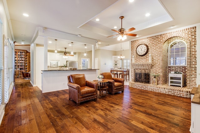 living room with ceiling fan with notable chandelier, dark hardwood / wood-style floors, ornamental molding, a brick fireplace, and a tray ceiling